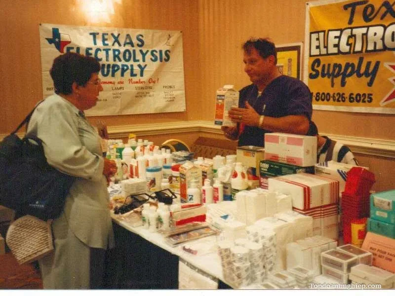 Vendor explaining products at a Texas Electrolysis Supply exhibition booth surrounded by treatment supplies