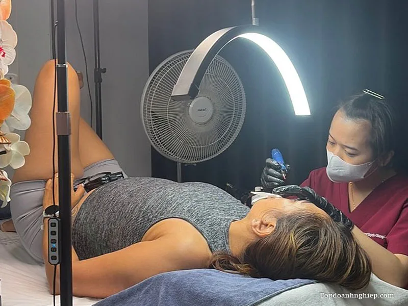 Specialist wearing black gloves performing delicate cosmetic tattooing under a curved LED light at VietBeautyHub