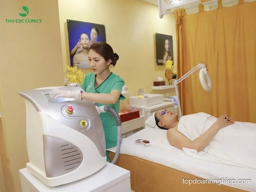 Technician in a green uniform calibrating an aesthetic skin therapy machine beside a client at Thu Cuc Clinics